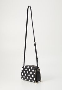 Sac crossbody noir avec des pois blancs, fermeture zippée, sangle réglable et accents en métal doré. Texture lisse et forme rectangulaire.