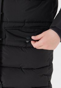 Sort pufferjakke med en synlig frontlomme, der har en trykknaplukning. Stoffet ser glat ud med en quilted struktur.