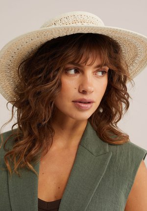 Femme aux cheveux bruns ondulés portant un chapeau tissé à large bord et un blazer vert sans manches, regardant vers sa droite sur un fond neutre.