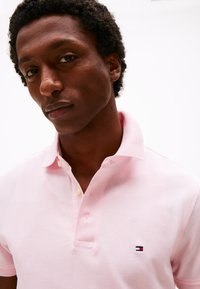 Világos rózsaszín, texturált pamutból készült galléros póló, kétgombos gallérral és egy kis hímzett logóval a bal mellkason.