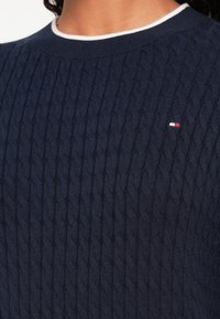 Tmavomodrý pletený sveter so vzorom vrkoča, bielym lemovaným okrúhlym golierom a malým červeno-bielo-modrým logom na hrudi.