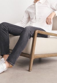 Jeans en denim gris foncé, coupe slim avec des coutures contrastantes, associés à une chemise blanche texturée à boutons, et des baskets blanches.