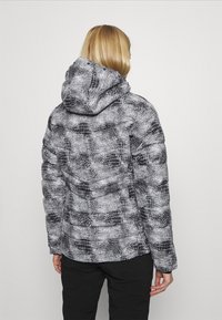 Kurtka puchowa w czarno-białym wzorze węża, z kapturem, pikowaną teksturą i elastycznymi mankietami. Noszona z czarnymi spodniami.