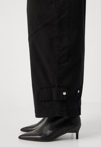 Stivali neri con tacco a stiletto e punta affusolata indossati con pantaloni neri che presentano dettagli con cinturini abbottonati vicino alle caviglie, su uno sfondo bianco.