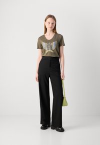 Olijfgroene graphic t-shirt met vleugeldesign, gecombineerd met zwarte wijde broek. Model houdt een kleine, gestructureerde, limoen groene handtas vast.
