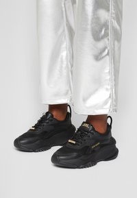 Sneakers neri con tomaia in mesh e pelle, dettagli dorati, punta arrotondata e suola spessa. Abbinati a pantaloni larghi metallici argento.