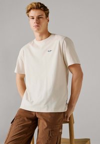 Ung mand med kort blondt hår iført en hvid kortærmet skjorte og brune cargobukser, der læner sig op ad en træstol foran en grå baggrund.