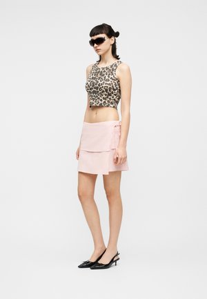 Kvinde iført leopardmønstret ærmeløs crop top, lyserød omslagsnederdel, sorte slingback-hæle og store sorte solbriller, stående i profil.