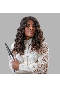 Trådløs hårkrølling med et elegant svart kropp og sølvdetaljer, holdt mot en teksturert hvit bluse med blonder som har blomster mønstre.