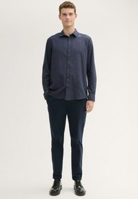 Camisa de manga larga azul marino con cuello abotonado, combinada con pantalones azul oscuro y zapatos negros, mostrada en una figura masculina de pie.