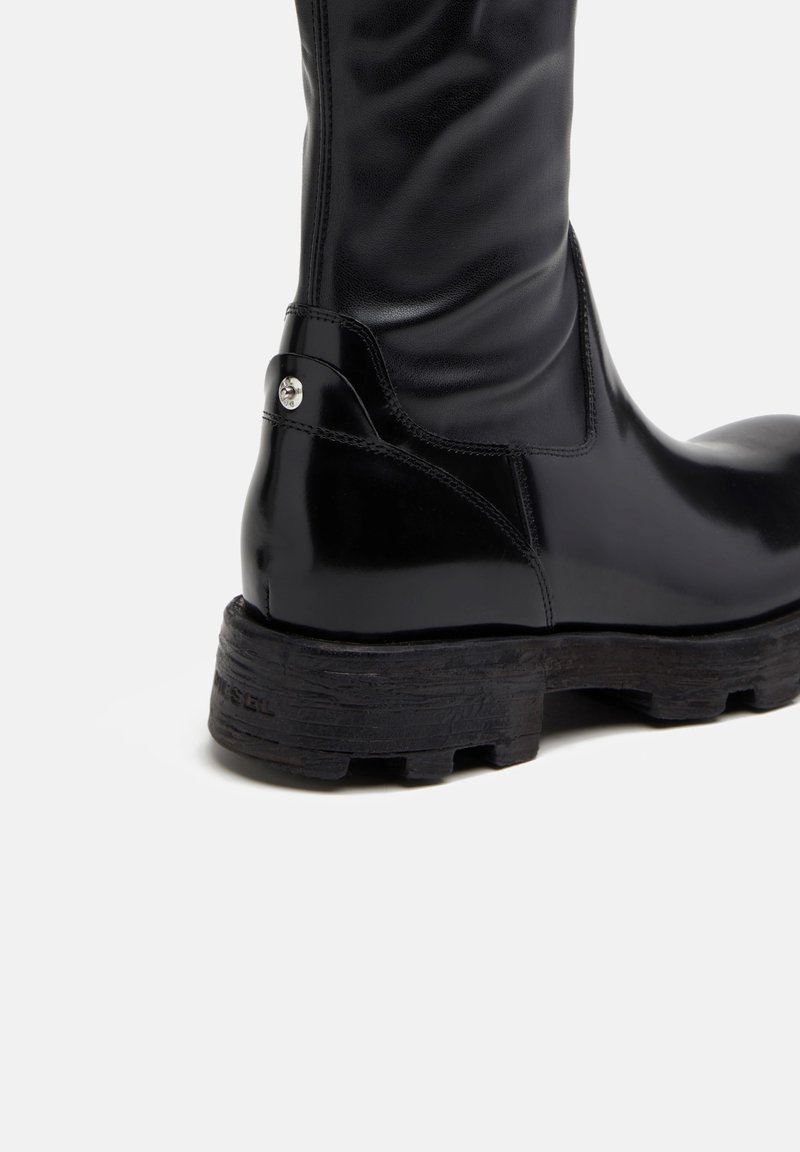 Bota negra hasta la rodilla con un upper de cuero brillante, suela de goma texturizada y un acento de botón plateado cerca del tobillo.