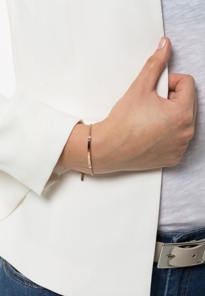Main posée sur la taille portant un bracelet chaîne en or rose avec une petite plaque rectangulaire, un blazer blanc, une chemise gris clair et un jean bleu.