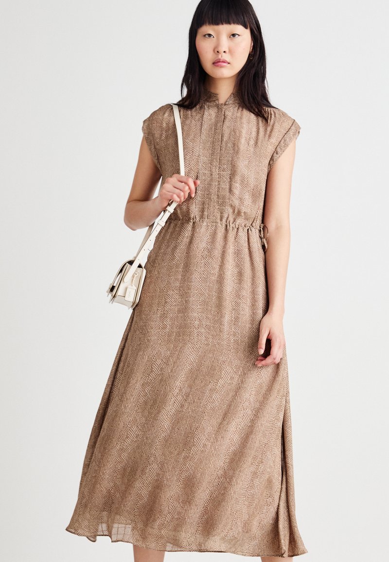 Rjava midi obleka s vzorcem, visokim ovratnikom, kratkimi rokavi s kapico in elastičnim pasom. Manekenka drži majhno belo torbico.