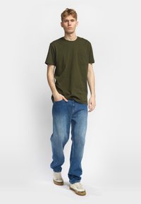 Camiseta de algodón verde oliva con bolsillo en el pecho, combinada con jeans de mezclilla azul claro. Modelo lleva zapatillas blancas con suelas de goma.