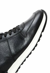 Zapatilla negra de cuero con una superficie texturizada, punta redonda, cordones negros y una suela de goma blanca con un patrón de huella negro.