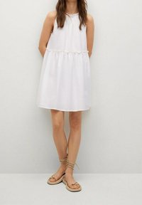 Jeune femme portant une robe sans manches blanche et des sandales plates beiges avec des brides à la cheville, se tenant contre un fond clair.