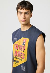 Marinblå ärmlös t-shirt med en stor gul grafik och röd text. Tillverkad av bomull, har den rund hals och drop shoulder-design.