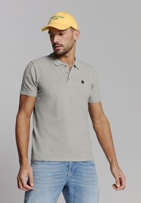 Grijze polo met een knoopsluiting van drie knopen en een klein logo op de borst, gecombineerd met een gele pet met groene tekst.