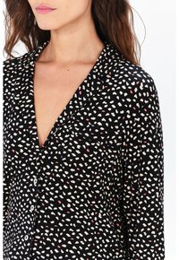 Blouse noire avec un col en V, ornée d'un motif dispersé de formes blanches et roses. Fermeture à boutons et texture de tissu douce.