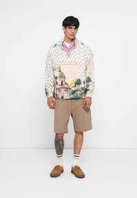 Lettvekts pulloverjakke i krem med et fargerikt landskapsprint. Brukt over en lavendel skjorte, sammen med beige plisserte shorts og brune sko.
