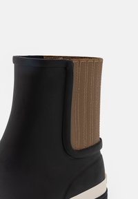 Černá gumová kotníková bota s hnědou žebrovanou elastickou stranou. Vybavená krémovou podrážkou pro kontrast. Hladký povrch se strukturovaným designem.