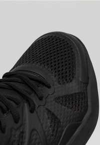 Chaussure de sport noire avec un dessus en mesh, semelle en caoutchouc texturé et lacets plats. Comprend des éléments de design découpés et une zone de renforcement au niveau des orteils.