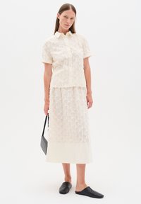 Cream-colored, embroidered top with short sleeves and button closure, paired with a matching long skirt. Black loafers and a small black bag.