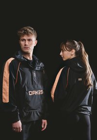 Mand og kvinde iført sorte hættejakker med ferskenfarvede striber og "OAK25" branding, poserer mod en sort baggrund.