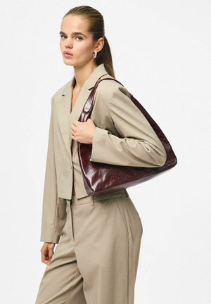 Nais beige ülikonnas, kes hoiab suurt tumedat pruuni õlakot, juuksed tagasi seotud ja kannab rõngas kõrvarõngaid.