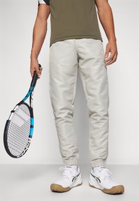 Pantalones atléticos color beige claro con puños elásticos, combinados con zapatillas blancas que tienen detalles en negro y dorado. Sujetando una raqueta de tenis negra y azul.