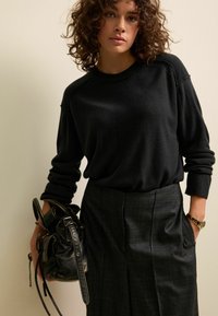 Maglione nero a maglia con scollatura rotonda, indossato con pantaloni grigi scozzesi sartoriali. Si tiene una borsa nera texturizzata, dotata di sottili manici.