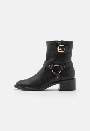 Bottine en cuir noir avec un petit talon carré, boucle en ton doré et sangle décorative avec accent circulaire, texture lisse.