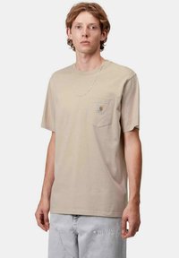 Camiseta de algodón color beige con mangas cortas, cuello redondo y bolsillo en el pecho izquierdo con una etiqueta. Complementada con unos jeans gris claro.