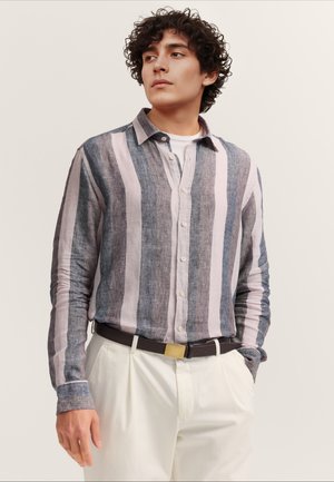 Junger Mann mit lockigem Haar, der ein langärmliges Hemd mit vertikalen Streifen in Blau, Grau und Weiß trägt, das in weiße Hosen mit dunklem Gürtel gesteckt ist.