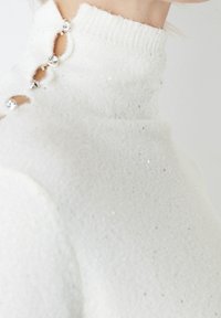 Suéter blanco de cuello alto hecho de una tela texturizada, con botones de rhinestones y sutiles detalles brillantes en todo su diseño.