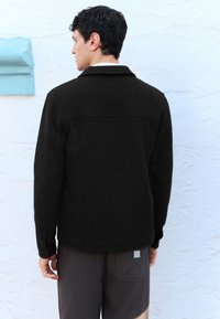 Melnas teksturētas jaka ar apkakli, kurai ir smalka rakstaina apdare. Dizainā ir iezīmēta etiķete uz muguras apakšmala un strukturētas piedurknes.