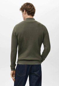 Jersey verde oliva con nervaduras, mangas largas y cuello alto, con un diseño ajustado y tela texturizada, combinado con vaqueros azul oscuro.