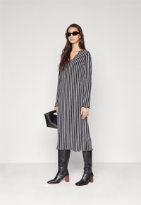 Robe longue rayée en noir et gris, avec un col en V, des manches longues et une silhouette fluide. Associée à des bottes noires ajustées jusqu'aux genoux.
