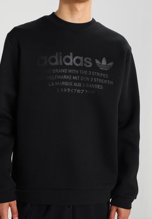 Personne portant un sweat-shirt Adidas noir avec le logo et un texte multilingue à propos de « La marque aux 3 bandes » sur la poitrine.