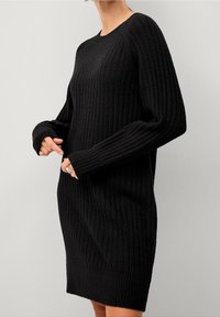 Robe-pull noire côtelée avec un col rond, des manches longues et une coupe décontractée. La texture est douce et légèrement extensible, avec un ourlet inférieur accentué.