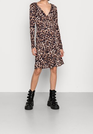 Kobieta w długiej sukience kopertowej w panterkę, w połączeniu z czarnymi botkami na kostkę z klamrą, stojąca na tle jednolitego jasnego tła.