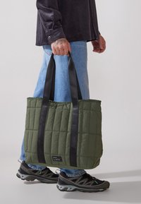 Grønn quilted toteveske med svarte stropper, med en teksturert overflate og et merke nederst, holdt av en person i denimjeans og sneakers.