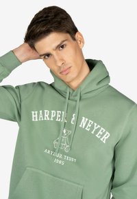 Sudadera verde con un bolsillo canguro, que presenta una impresión gráfica de un oso de peluche y el texto "HARPER & NEYER" en blanco. Material suave y corte relajado.