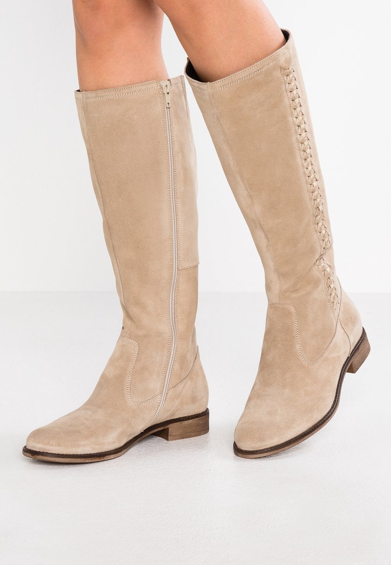 Bottes hautes jusqu'au genou en daim beige avec fermeture éclair latérale et détails tressés sur la tige extérieure. Talon plat, bout arrondi et texture lisse.