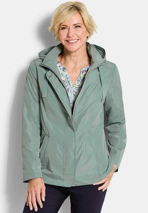 Lachende vrouw van middelbare leeftijd met kort blond haar, gekleed in een lichtgroene capuchonjas over een gebloesd blouse, staand tegen een witte achtergrond.