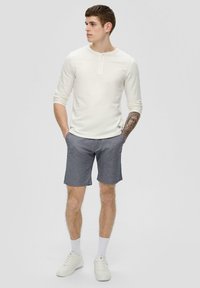 Weißes Langarm-Henley-Shirt mit Knopfleiste, kombiniert mit grauen Shorts. Das Model trägt weiße Sneaker und socken bis zur Knöchelhöhe in Weiß.