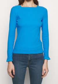 Blå långärmad topp med fyrkantig halsringning och figurnära design. Har volangärmar och ett mjukt, stretchigt bomullsmaterial. Kombineras med blå jeans.