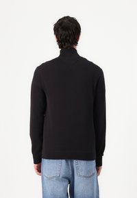 Pull en maille noire avec un col montant côtelé, des poignets côtelés et un ourlet côtelé. Présente un design simple sans motifs ni accents supplémentaires.