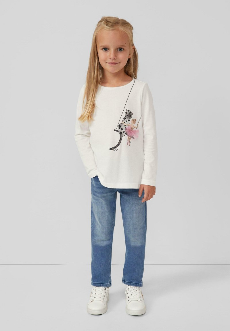 Camicia bianca a maniche lunghe con un design di un gatto giocoso, jeans azzurri e sneakers bianche. La camicia presenta un dettaglio di tutù rosa nella grafica.