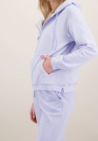 Lila mikina na zip z měkké látky. Má přední kapsu, stahovací šňůrky a žebrované manžety. Doplněna o odpovídající kalhoty.